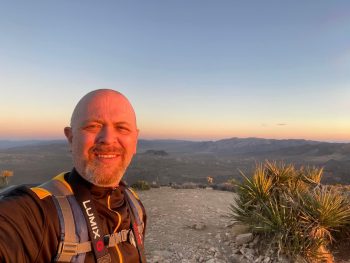 A journey unfolds as a person in hiking gear stands on a rocky path at sunset, with a vast desert landscape and distant mountains stretching endlessly in the background.