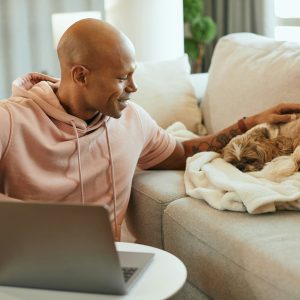A man sitting on a couch with a laptop pets one of his shih tzus, who is sleeping peacefully on a blanket.
