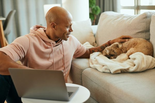 A man sitting on a couch with a laptop pets one of his shih tzus, who is sleeping peacefully on a blanket.