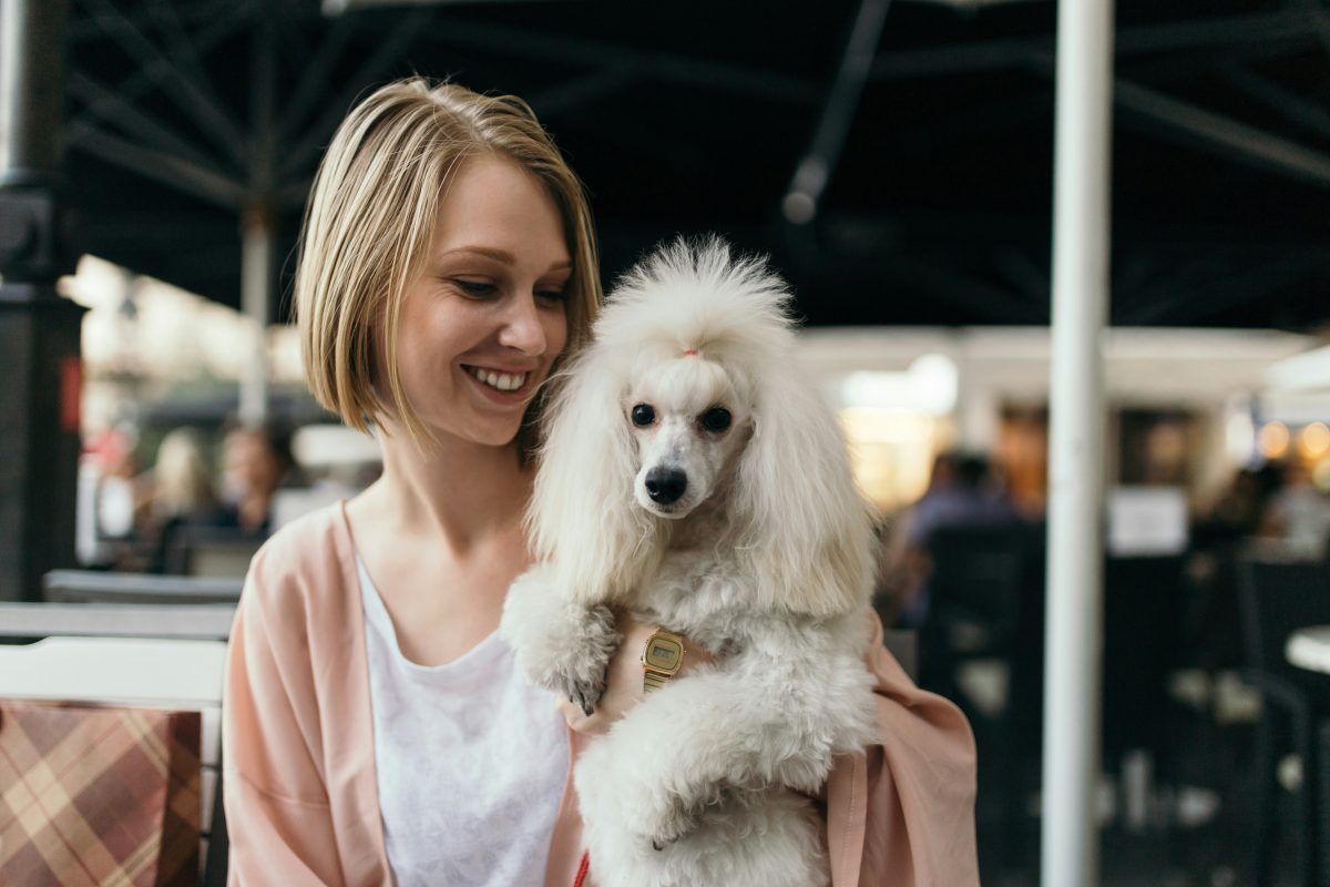 A woman with short blond hair smiles while holding a fluffy poodle in her arms at an outdoor setting.