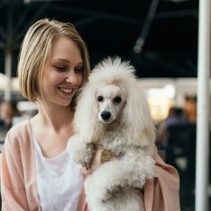 A woman with short blond hair smiles while holding a fluffy poodle in her arms at an outdoor setting.