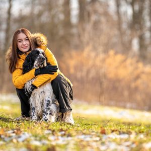 A woman in a yellow coat crouches on grass, hugging a black and white dog. Trees in the background suggest a winter setting.