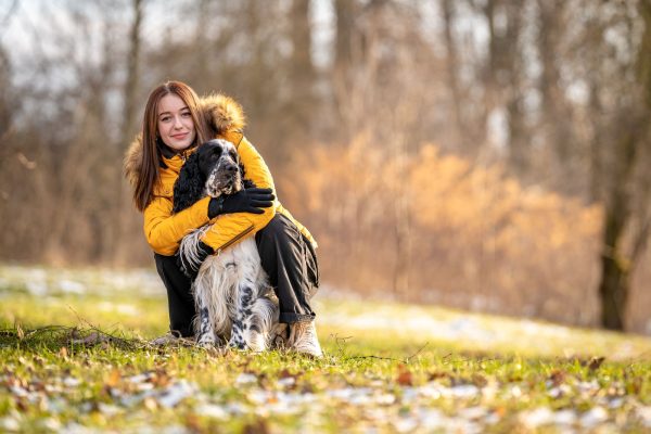 A woman in a yellow coat crouches on grass, hugging a black and white dog. Trees in the background suggest a winter setting.