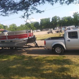 A silver pickup truck towing a red pontoon boat on a trailer is parked on a grassy area, perhaps preparing for another dinghy shipping adventure. Trees and parked vehicles form the backdrop of this scene.