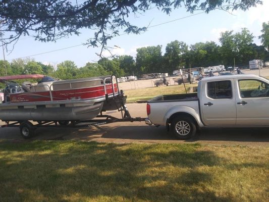 A silver pickup truck towing a red pontoon boat on a trailer is parked on a grassy area, perhaps preparing for another dinghy shipping adventure. Trees and parked vehicles form the backdrop of this scene.