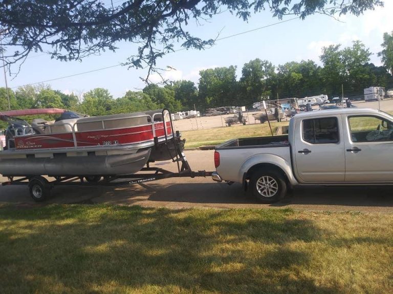 A silver pickup truck towing a red pontoon boat on a trailer is parked on a grassy area, perhaps preparing for another dinghy shipping adventure. Trees and parked vehicles form the backdrop of this scene.