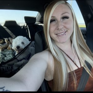 Josie Trista Norwood, embracing life on the road, snaps a front-seat selfie. Her long blonde hair frames her face, while her small white dog relaxes in the back seat beside a stuffed giraffe and blanket.