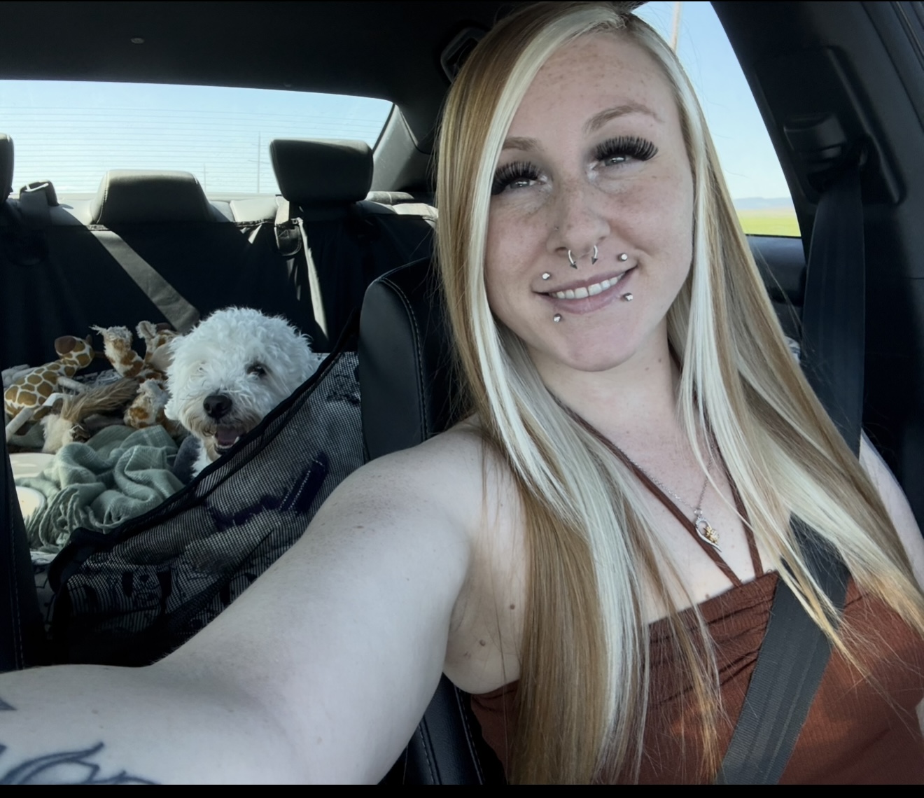 Josie Trista Norwood, embracing life on the road, snaps a front-seat selfie. Her long blonde hair frames her face, while her small white dog relaxes in the back seat beside a stuffed giraffe and blanket.