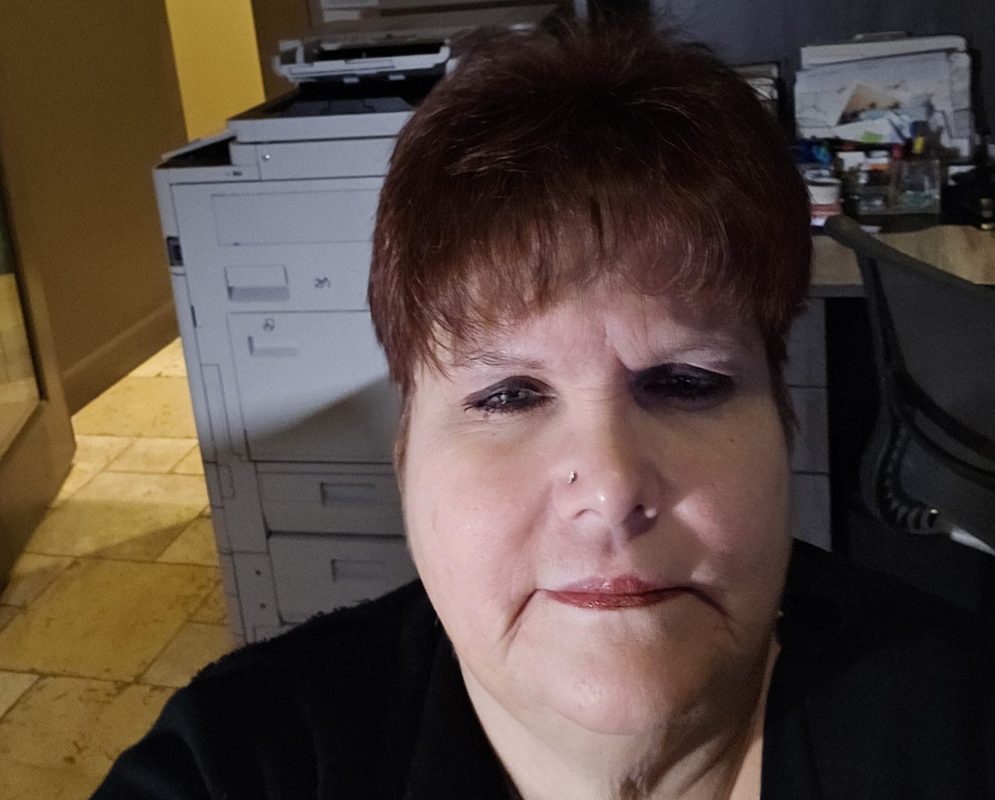 A woman with short brown hair and a nose piercing, possibly a CitizenShipper driver like Shery Mathewson, is sitting indoors facing the camera, with office equipment and shelves visible in the background.