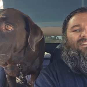A bearded man smiles at the camera next to a large brown dog sitting in the backseat of a car. This pet transporter captures a cozy moment as his furry passenger looks out the window while he’s partially in the frame on the right side.
