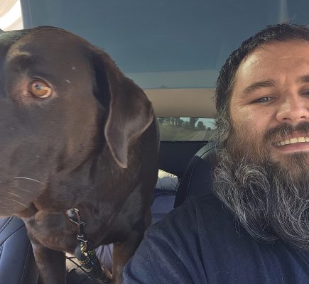 A bearded man smiles at the camera next to a large brown dog sitting in the backseat of a car. This pet transporter captures a cozy moment as his furry passenger looks out the window while he’s partially in the frame on the right side.
