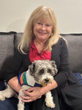 Dr. Neely with long blonde hair sits on a couch, wearing a red shirt and black jacket. She is holding a small white and gray dog in a colorful striped sweater, caring for her furry friend’s pet health with attention and love.