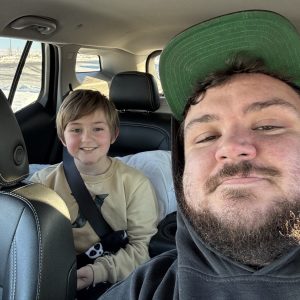 A man in a green cap and black hoodie sits in the front seat of a car, smiling for a selfie as if ready for a transporter interview. A child with light brown hair and a beige sweater sits in the back, also smiling, holding a game controller.