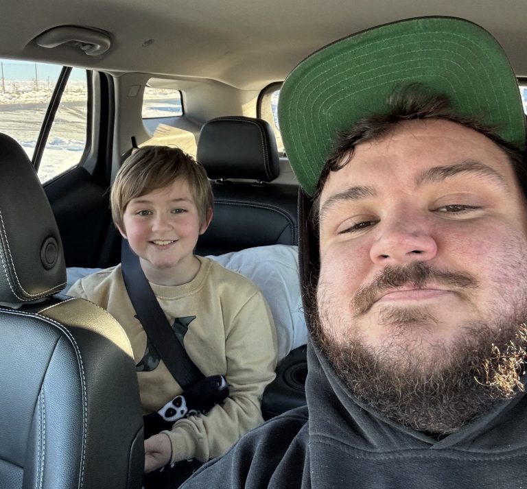 A man in a green cap and black hoodie sits in the front seat of a car, smiling for a selfie as if ready for a transporter interview. A child with light brown hair and a beige sweater sits in the back, also smiling, holding a game controller.