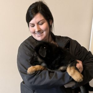 A woman with dark hair smiles while holding a fluffy black and brown puppy in her arms, standing indoors against a light-colored wall—showcasing her passion for animal care.