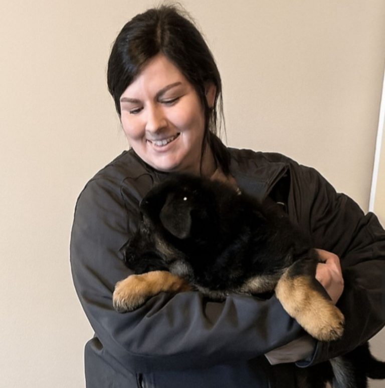 A woman with dark hair smiles while holding a fluffy black and brown puppy in her arms, standing indoors against a light-colored wall—showcasing her passion for animal care.