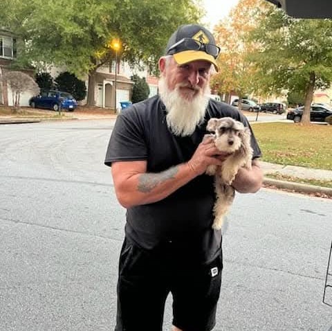 An older man with a white beard, sunglasses on his cap, and a black t-shirt holds a small gray and white dog during a transporter interview on a residential street lined with houses and trees.