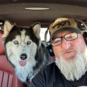 A man with a white beard, glasses, headphones, and a cap takes a selfie in a car during his transporter interview. Next to him, a husky dog with blue eyes and its tongue out sits on the red leather seat.