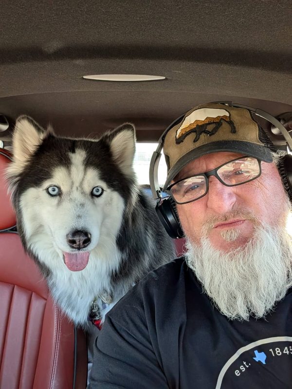 A man with a white beard, glasses, headphones, and a cap takes a selfie in a car during his transporter interview. Next to him, a husky dog with blue eyes and its tongue out sits on the red leather seat.