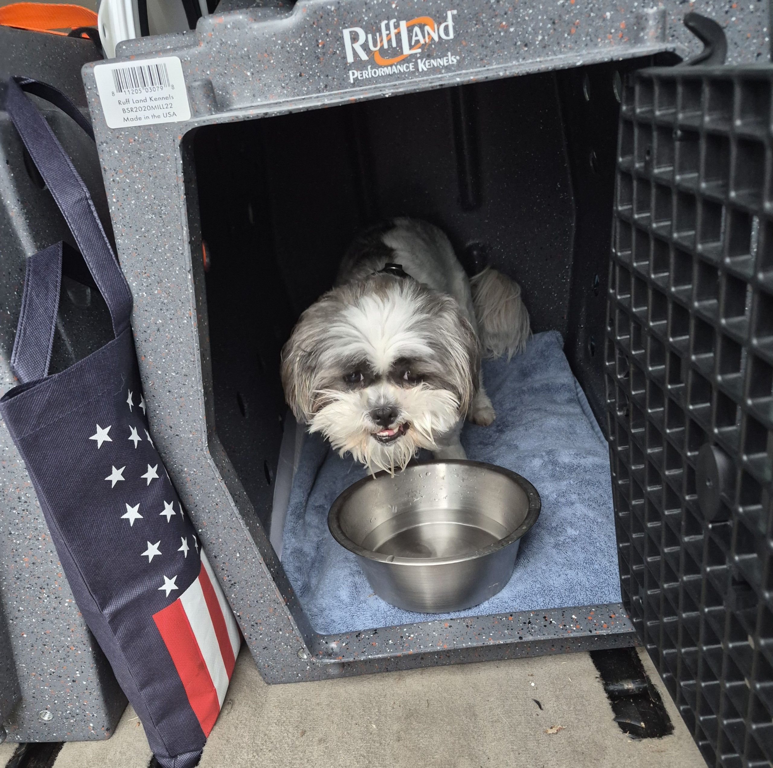 A small, fluffy black and white dog sits inside a RuffLand kennel on a blue towel, with a metal water bowl in front—just one of many in a van full of dogs. An American flag bag hangs on the side of the kennel.