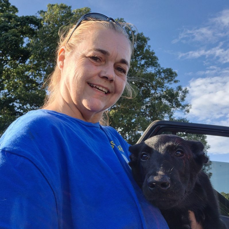 A smiling woman in a blue shirt stands outdoors, holding a black dog close to her chest. Green trees and a partly cloudy sky frame Tammy Leopard, who enjoys a break from corporate life with her furry companion.