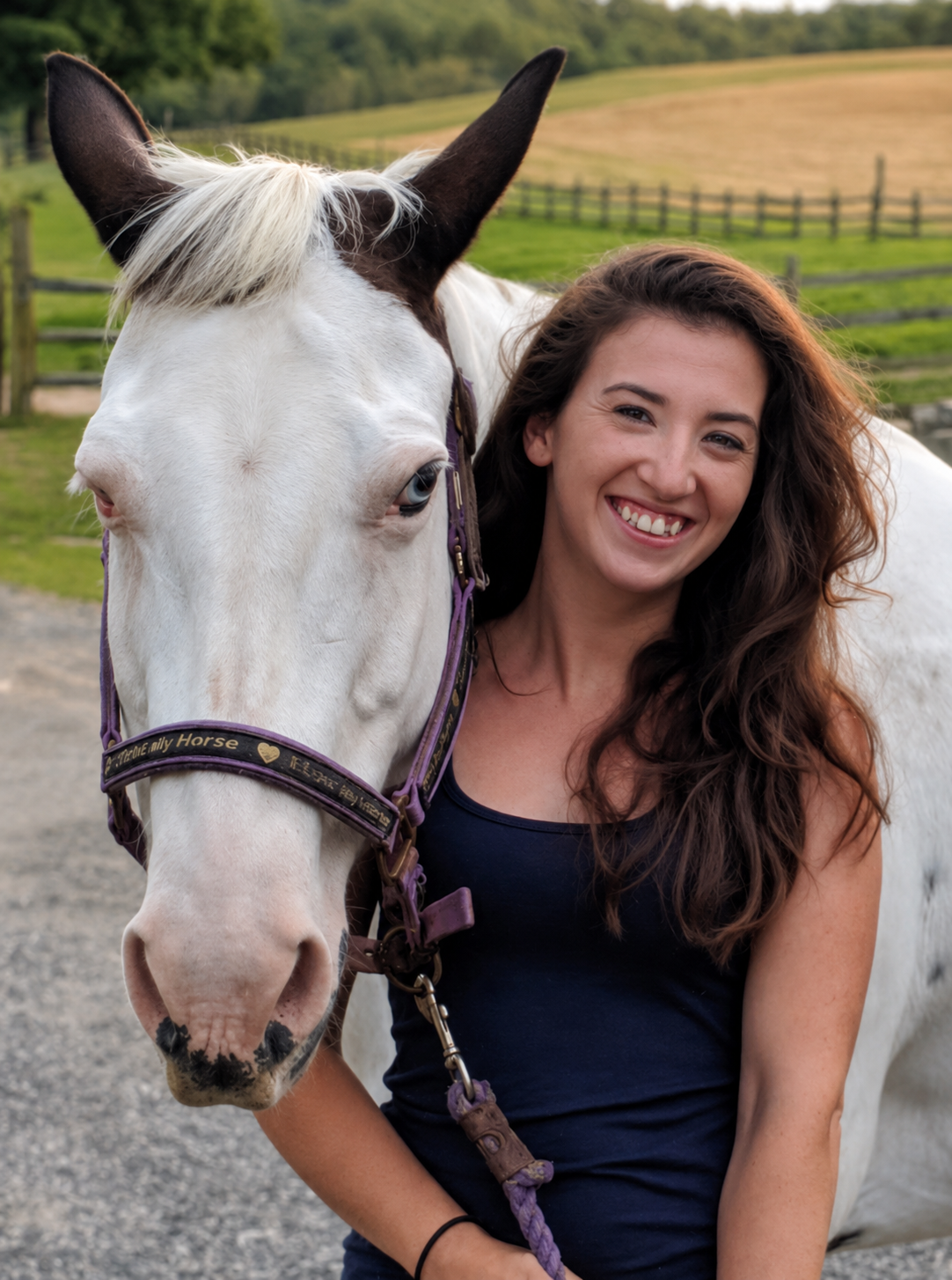 A woman smiling and standing beside a white horse with dark ears and a halter, outdoors near a fence and grassy field—an ideal moment for anyone seeking a friendly horse hauler or reliable pet transporter.