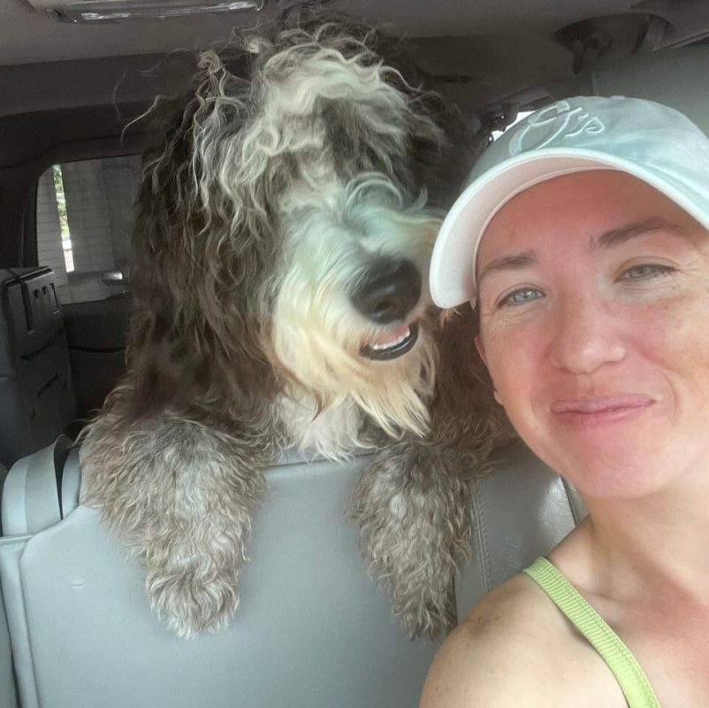 A woman in a light gray cap smiles for a selfie inside a car, with a fluffy, curly-haired dog leaning over the back seat beside her—capturing a joyful moment during her journey as a dedicated pet transporter.