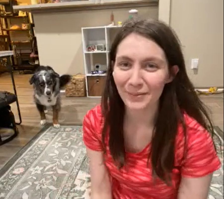 A woman with long brown hair in a red shirt sits on a patterned rug indoors, smiling at the camera. Behind her, a dog with black, white, and brown fur—perhaps an adorable Mini Aussie awaiting transport via CitizenShipper from DFW—stands near a shelf.